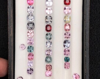 May include: A collection of loose, cushion-cut gemstones arranged in three vertical rows within a white display tray. The stones feature a variety of colours including pink, red, blue, green, yellow, and clear, with two additional stones placed outside the rows.