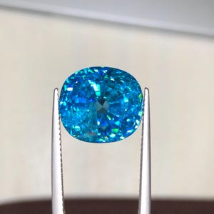 May include: A vibrant, oval-cut gemstone, held by silver tweezers. The gem displays a brilliant, multifaceted surface with shades of turquoise and blue, reflecting light. The gemstone's clarity and color are the focal point.