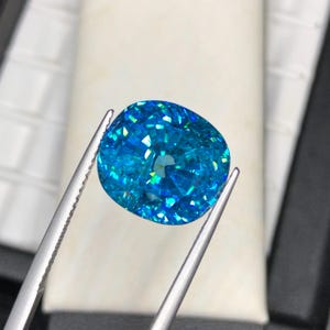 May include: A brilliant, cushion-cut gemstone in shades of blue and turquoise is held by silver tweezers. The gem's facets reflect light, creating a sparkling effect. The background is a soft, neutral color.