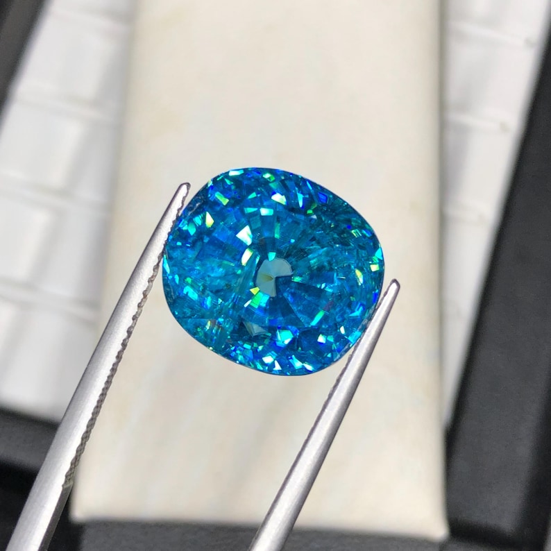 May include: A brilliant, cushion-cut gemstone in shades of blue and turquoise is held by silver tweezers. The gem's facets reflect light, creating a sparkling effect. The background is a soft, neutral color.