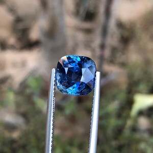 May include: A square-cut, deep blue gemstone is held by silver tweezers. The gem's facets reflect light, creating a sparkling effect. The background is blurred, with hints of green and brown.