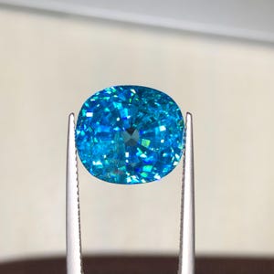 May include: A vibrant, oval-cut gemstone held by silver tweezers. The gem is a brilliant shade of blue with internal facets that reflect light, creating a sparkling effect. The background is a soft, neutral tone, highlighting the gem's color and clarity.