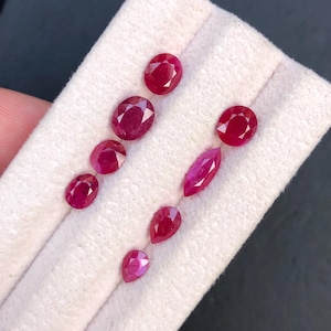 Puede incluir: Colección de rubíes facetados de formas ovaladas y de pera, de varios tamaños. Las piedras preciosas son de color rojo intenso y se muestran sobre una tela blanca. La disposición resalta los diferentes cortes y el brillo de los rubíes.