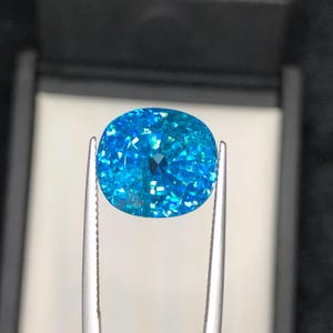 May include: A brilliant, cushion-cut gemstone in shades of turquoise and aqua is held by silver tweezers. The gem's facets reflect light, creating a sparkling effect. The background is a dark, neutral tone, highlighting the gem's vibrant color.