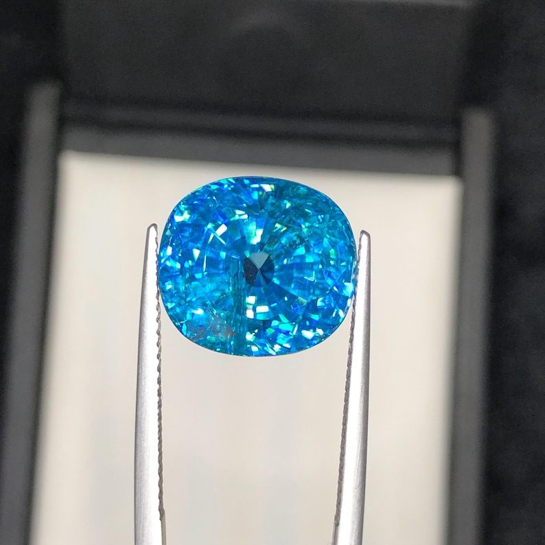 May include: A brilliant, cushion-cut gemstone in shades of turquoise and aqua is held by silver tweezers. The gem's facets reflect light, creating a sparkling effect. The background is a dark, neutral tone, highlighting the gem's vibrant color.