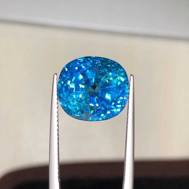 May include: A brilliant, oval-cut gemstone held by tweezers. The gem is a vibrant, sparkling blue with internal facets that reflect light. The overall shape is symmetrical, and the gem appears to be of high quality.