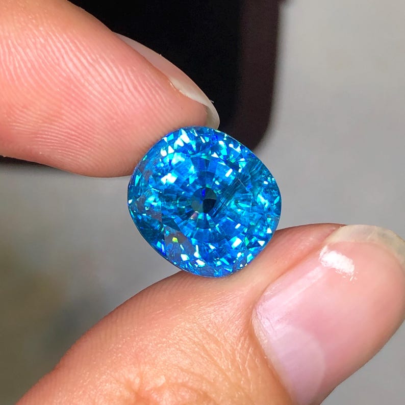 May include: A vibrant, cushion-cut, blue gemstone held between fingers. The gem's facets reflect light, creating a sparkling effect. The color is a bright, clear blue, with a slightly rounded square shape. The gemstone is the focal point of the image.