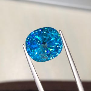 May include: A brilliant, oval-cut gemstone with a vibrant, light blue hue. The gem is held by silver tweezers, showcasing its facets and internal reflections. The gemstone's color shifts with light, displaying flashes of green and blue.