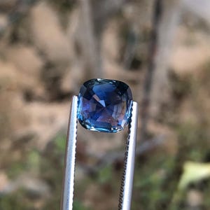 May include: A square-cut gemstone, held by tweezers, exhibits a rich blue colour with flashes of purple and turquoise. The cut facets catch the light, producing a shimmering effect. The blurred background highlights the gem's clarity and colour.