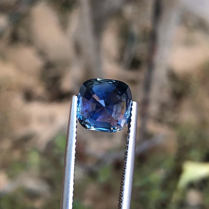 May include: A square-cut gemstone, held by tweezers, exhibits a rich blue colour with flashes of purple and turquoise. The cut facets catch the light, producing a shimmering effect. The blurred background highlights the gem's clarity and colour.