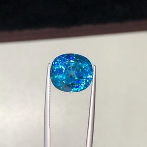 May include: A vibrant, oval-cut gemstone, held by silver tweezers. The gem is a brilliant shade of blue with internal facets that reflect light, creating a sparkling effect. The background is a neutral tone, emphasizing the gem's color and clarity.