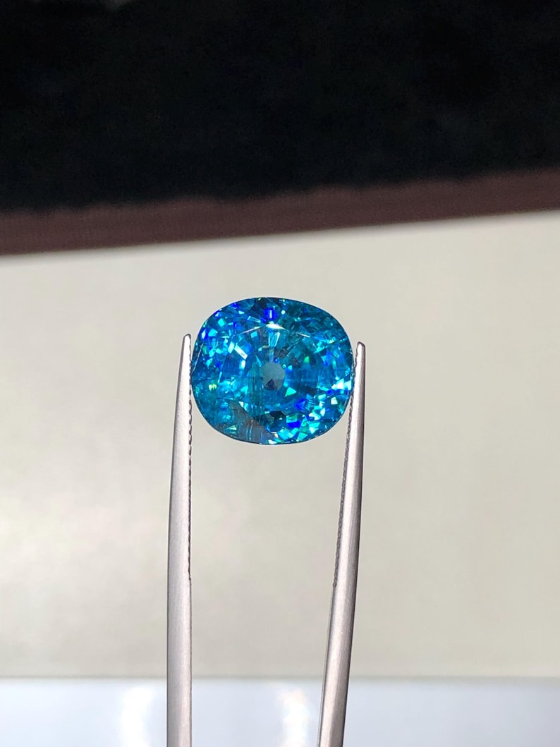 May include: A vibrant, oval-cut gemstone, held by silver tweezers. The gem is a brilliant shade of blue with internal facets that reflect light, creating a sparkling effect. The background is a neutral tone, emphasizing the gem's color and clarity.