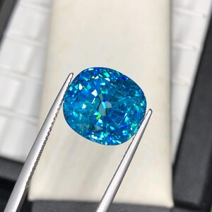 May include: A large, cushion-cut gemstone with a vibrant, light blue hue. The gem is held by silver tweezers, showcasing its facets and internal reflections. The stone's surface sparkles with various shades of blue and green.
