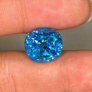 May include: A brilliant, cushion-cut gemstone with a vibrant, sparkling blue hue. The facets reflect light, creating a dazzling display of color and brilliance. The gemstone is held between two fingers.