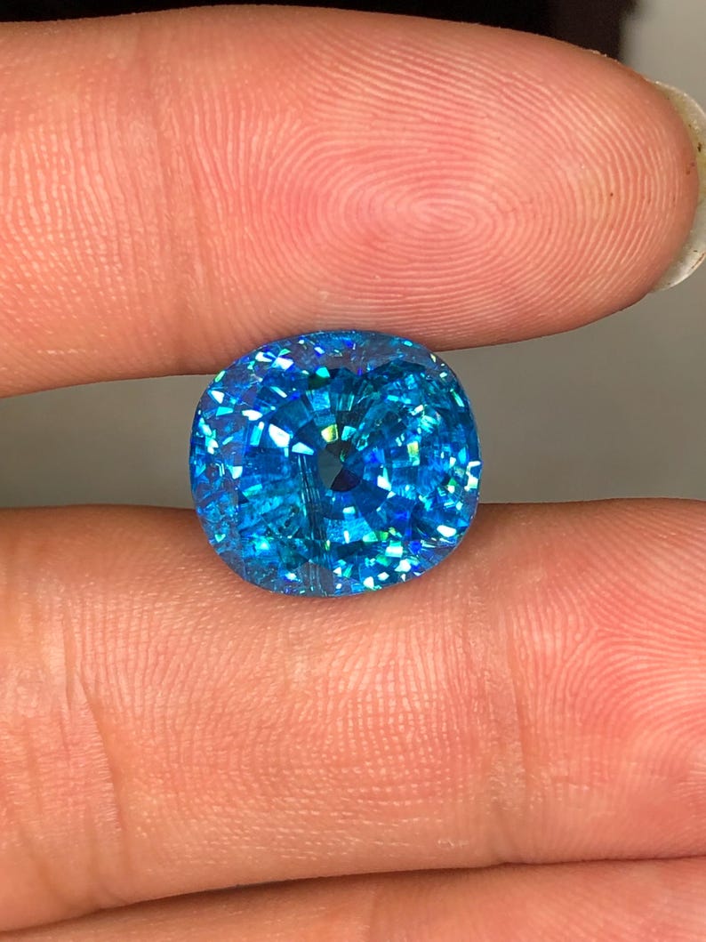 May include: A brilliant, cushion-cut gemstone with a vibrant, sparkling blue hue. The facets reflect light, creating a dazzling display of color and brilliance. The gemstone is held between two fingers.