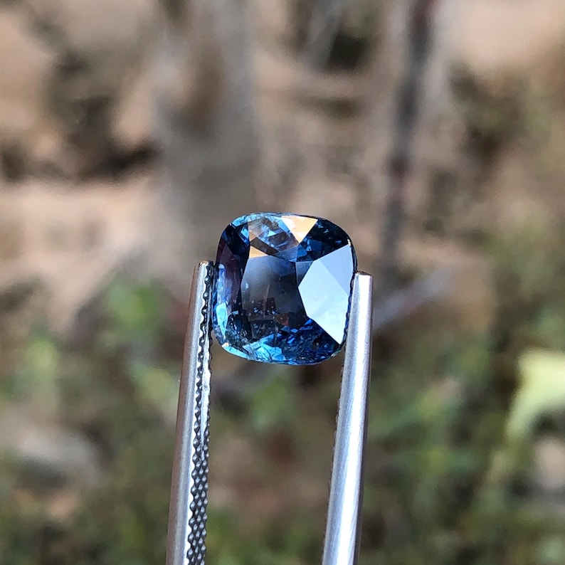 May include: A square-cut, blue gemstone held by silver tweezers. The gem's facets reflect light, displaying internal patterns. The background is blurred, suggesting an outdoor setting. The gem is likely measured in millimeters.