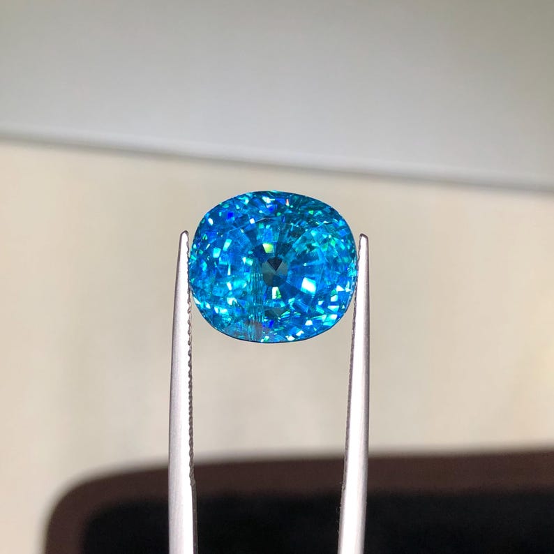 May include: A vibrant, cushion-cut gemstone held by silver tweezers. The gem is a brilliant shade of turquoise blue, with facets that catch and reflect light. The shape is oval, and the gem appears to be flawless.