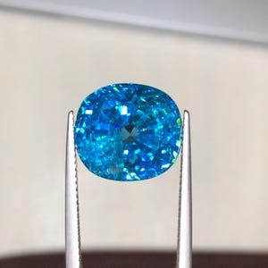 May include: A brilliant, oval-cut gemstone held by silver tweezers. The gem is a vibrant, sparkling blue with facets that catch the light, creating a dazzling display of color and brilliance. The gem's shape is symmetrical, and the tweezers provide a neutral background.