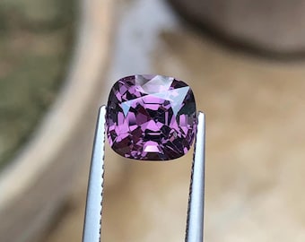 May include: A cushion-cut, faceted gemstone in shades of purple is held by tweezers. The gem has a high degree of clarity, with multiple facets reflecting light. The gemstone's color ranges from a light lavender to a deep violet.