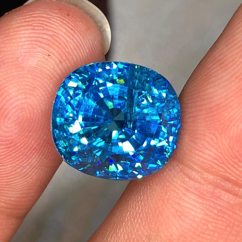 May include: A brilliant, round-cut gemstone with a vibrant, sparkling blue hue. The gem is held between two fingers, showcasing its facets and internal reflections. The stone's color ranges from light to deep blue, with flashes of green and white.