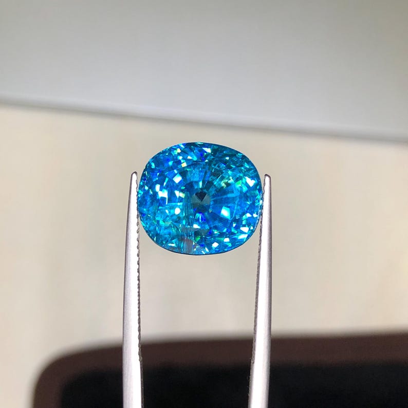 May include: A brilliant, oval-cut gemstone held by tweezers. The gem is a vibrant, sparkling blue with internal facets that reflect light. The background is a soft, neutral tone, highlighting the gem's color and clarity.
