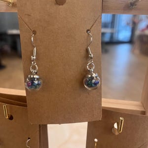 May include: Silver-toned dangle earrings. Each earring features a small, clear glass orb filled with colorful beads. The earrings are displayed on a brown cardboard earring display.