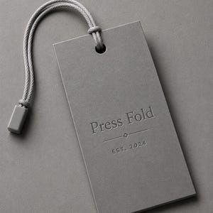 Puede incluir: Una etiqueta rectangular de papel gris con un cord&oacute;n gris y un cierre de pl&aacute;stico. La etiqueta presenta el texto en relieve Press Fold, un peque&ntilde;o icono de diamante y el texto EST. 2024 centrados en la parte frontal con una fuente clara.