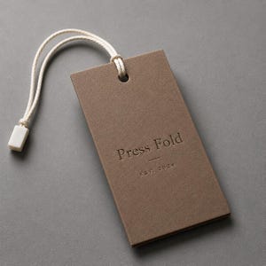 Puede incluir: Una etiqueta colgante rectangular de papel marr&oacute;n con el texto en relieve Press Fold y EST. 2024. La etiqueta incluye un lazo de cuerda blanca con un peque&ntilde;o cierre de pl&aacute;stico blanco sujeto a trav&eacute;s de un agujero en la parte superior.