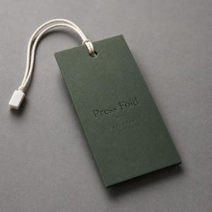 Puede incluir: Una etiqueta colgante rectangular de papel verde oscuro con un cord&oacute;n blanco y un cierre de pl&aacute;stico blanco. La etiqueta presenta el texto en relieve Press Fold y el texto m&aacute;s peque&ntilde;o EST. 2024 centrados en la parte frontal en letra sans-serif.