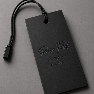 Puede incluir: Una etiqueta colgante rectangular negra con un cord&oacute;n negro y un cierre de pl&aacute;stico negro. La etiqueta presenta el texto Press Fold en relieve con una fuente manuscrita, un peque&ntilde;o gr&aacute;fico decorativo de hoja y el texto EST. 2024 en una fuente sans-serif.