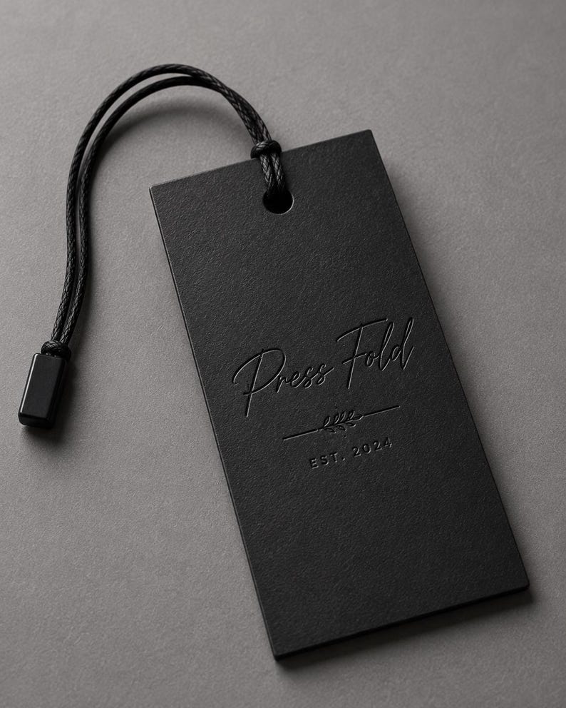 Puede incluir: Una etiqueta colgante rectangular negra con un cord&oacute;n negro y un cierre de pl&aacute;stico negro. La etiqueta presenta el texto Press Fold en relieve con una fuente manuscrita, un peque&ntilde;o gr&aacute;fico decorativo de hoja y el texto EST. 2024 en una fuente sans-serif.