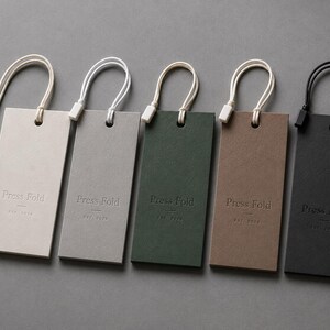 Puede incluir: Cinco etiquetas colgantes rectangulares de papel en color crema, gris claro, verde oscuro, marr&oacute;n y negro. Cada etiqueta presenta el texto en relieve Press Fold y est. 2023. Est&aacute;n unidas a cordones con cierres de pl&aacute;stico a juego.