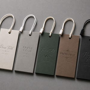 Puede incluir: Cinco etiquetas colgantes rectangulares de papel en beige, gris claro, verde oscuro, marr&oacute;n y negro. Cada etiqueta presenta el texto en relieve Press Fold y EST. 2024. Las etiquetas est&aacute;n unidas a cordones blancos o negros con cierres.
