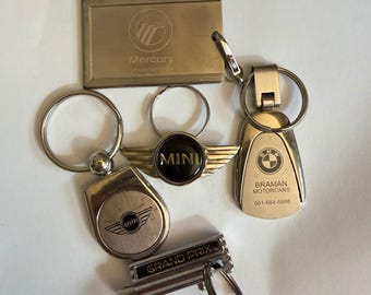 May include: A collection of five metal keychains featuring various automotive logos. Included are a rectangular Mercury tag, a circular Mini logo, a winged Mini emblem, a BMW Braman Motorcars fob with phone number, and a Grand Prix branded keychain.