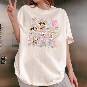 May include: Cream-colored t-shirt featuring Disney characters Minnie Mouse, Mickey Mouse, Donald Duck, Daisy Duck, and Pluto, each framed in a star shape. The word "magical" is printed below the characters in pastel colors. Pink bows and balloons complete the design.