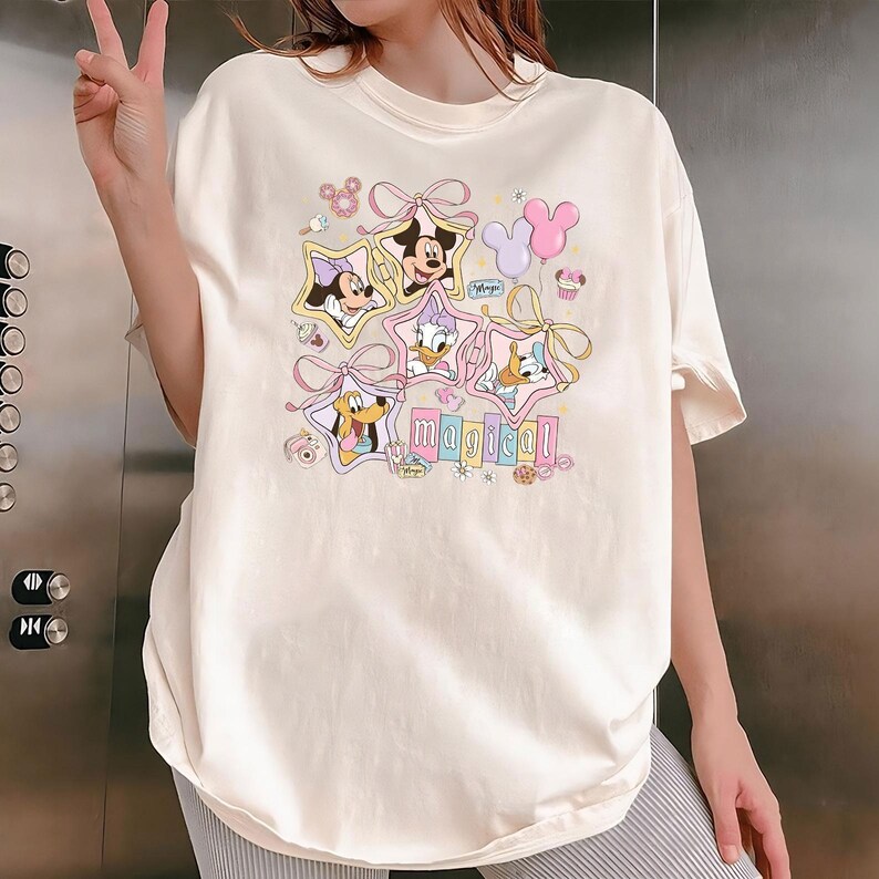 May include: Cream-colored t-shirt featuring Disney characters Minnie Mouse, Mickey Mouse, Donald Duck, Daisy Duck, and Pluto, each framed in a star shape. The word "magical" is printed below the characters in pastel colors. Pink bows and balloons complete the design.