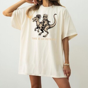 Op de afbeelding: Cr&egrave;mekleurig oversized T-shirt met een zwart-witte afbeelding van een kat met een kroon die op een dinosaurus rijdt. De tekst "THIS IS AN OUTRAGE" staat eronder. Het shirt heeft korte mouwen en een relaxte pasvorm.