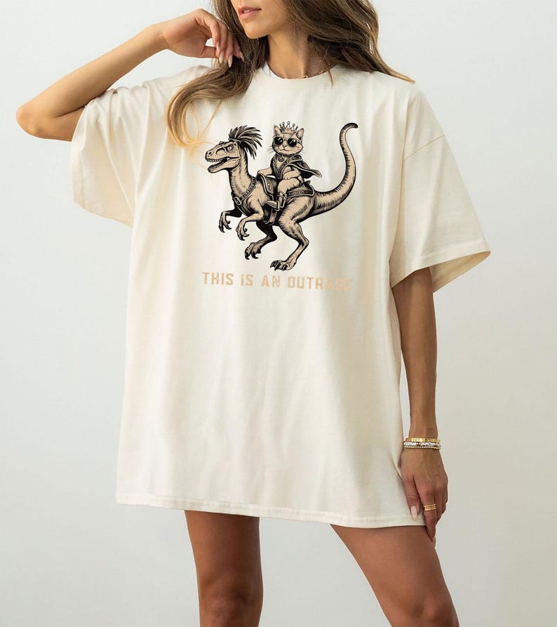 Op de afbeelding: Cr&egrave;mekleurig oversized T-shirt met een zwart-witte afbeelding van een kat met een kroon die op een dinosaurus rijdt. De tekst "THIS IS AN OUTRAGE" staat eronder. Het shirt heeft korte mouwen en een relaxte pasvorm.