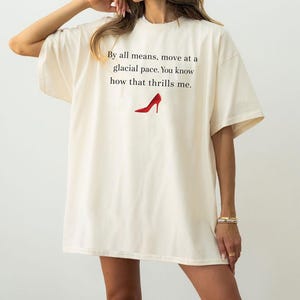 May include: Oversized cream t-shirt featuring the text "By all means, move at a glacial pace. You know how that thrills me." and a red high-heeled shoe graphic. The shirt has short sleeves and a loose fit.
