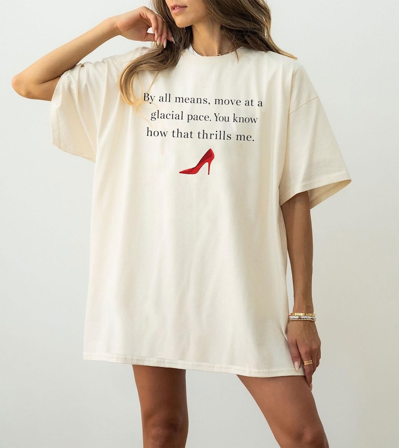 May include: Oversized cream t-shirt featuring the text "By all means, move at a glacial pace. You know how that thrills me." and a red high-heeled shoe graphic. The shirt has short sleeves and a loose fit.