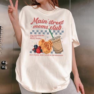 May include: Cream-colored t-shirt with the text "Main street moms club" in red and blue. The graphic includes a coffee drink, Mickey waffles, and berries. The shirt also has the text "Stop and smell the Coffee & Mickey Waffles" and "BUT, PLEASE, DON'T STOP IN THE WALKWAY."