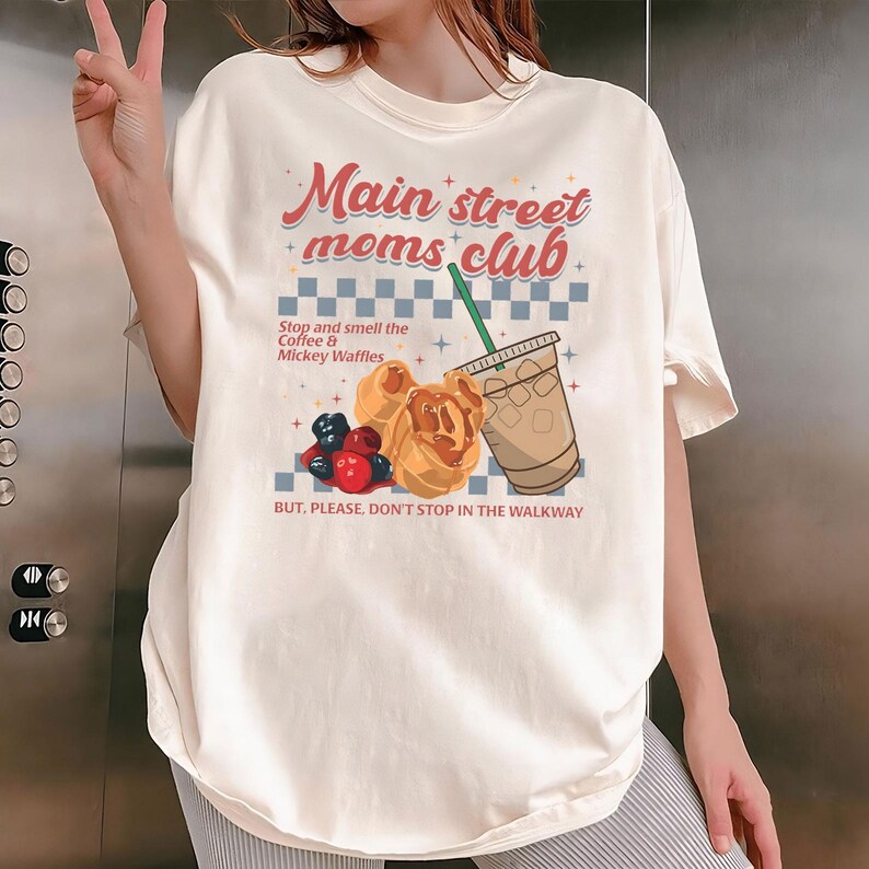 May include: Cream-colored t-shirt with the text "Main street moms club" in red and blue. The graphic includes a coffee drink, Mickey waffles, and berries. The shirt also has the text "Stop and smell the Coffee & Mickey Waffles" and "BUT, PLEASE, DON'T STOP IN THE WALKWAY."