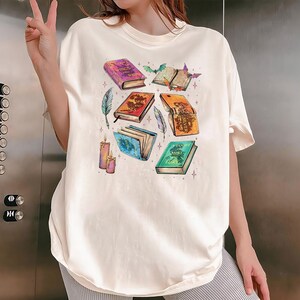 May include: Cream-colored t-shirt featuring a collection of illustrated books with titles like "Wings of Ruin" and "Silver Flame." The design includes feathers, candles, and bats, creating a fantasy theme. The shirt has a relaxed fit.