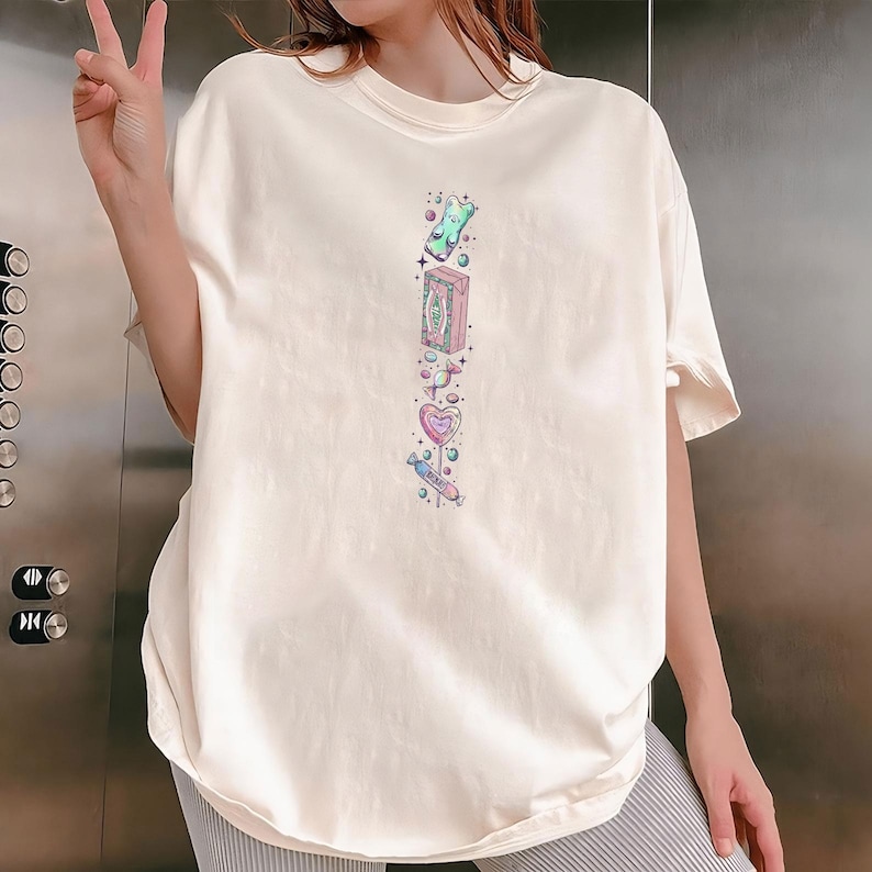 Puede incluir: Camiseta color crema con un dise&ntilde;o vertical de acuarela de caramelos y dulces. El dise&ntilde;o incluye un osito de goma, una caja, una piruleta en forma de coraz&oacute;n y un bast&oacute;n de caramelo. La camiseta es de corte holgado.