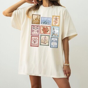 May include: Cream-colored oversized t-shirt featuring nine postage stamp-style graphics. The stamps display various illustrations and text, including "Lunathion," "The Night Court," and "Fireheart." The shirt has short sleeves and a relaxed fit.