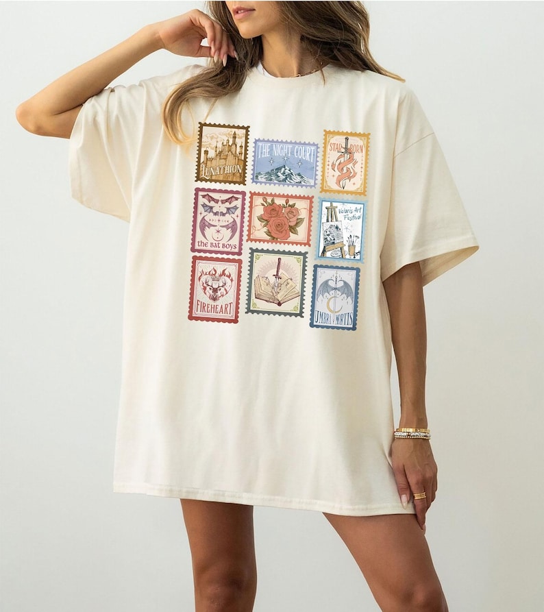 May include: Cream-colored oversized t-shirt featuring nine postage stamp-style graphics. The stamps display various illustrations and text, including "Lunathion," "The Night Court," and "Fireheart." The shirt has short sleeves and a relaxed fit.