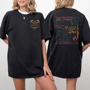 May include: Black t-shirt with a gold graphic on the front and text on the back. The front graphic features a mountain scene with a crescent moon and stars. The back of the shirt has the text "HELLO FEYRE DARLING" and other phrases.