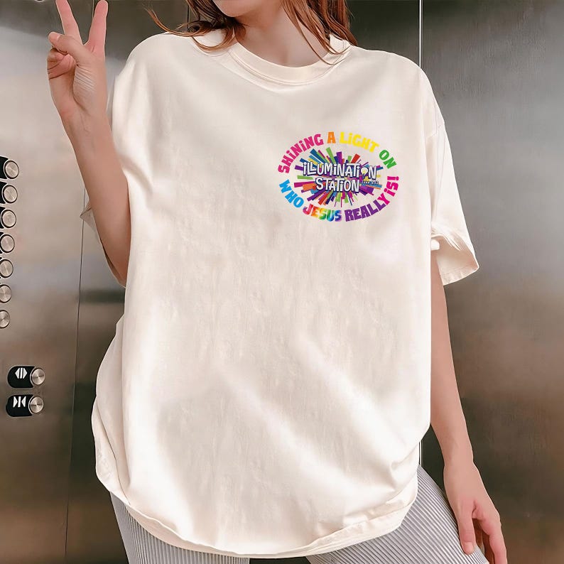 May include: Cream-colored t-shirt with a colorful circular graphic. The graphic reads "Shining a Light on Illumination Station Who Jesus Really Is" in a rainbow of colors. The shirt has short sleeves and a relaxed fit.