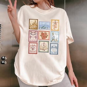 May include: Cream-colored t-shirt featuring nine postage stamp-style designs. Each stamp has a unique illustration and text, including a castle, mountains, roses, and the words "Fireheart" and "Umbra Mortis".
