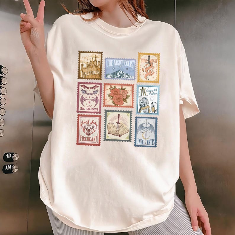 May include: Cream-colored t-shirt featuring nine postage stamp-style designs. Each stamp has a unique illustration and text, including a castle, mountains, roses, and the words "Fireheart" and "Umbra Mortis".